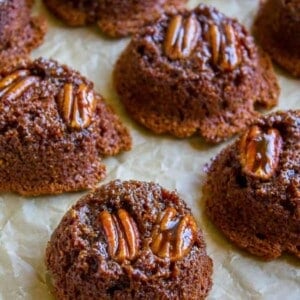Bran muffins topped with glossy pecan halves are arranged on parchment paper. Each mini cake features a rich, moist texture and a decorative, appetizing pecan on top.