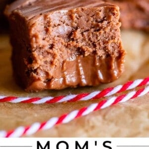 A close-up of a thick, creamy chocolate fudge square with a bite taken out, resting on parchment paper, rich with marshmallow flavor. More fudge pieces are blurred in the background. The text at the bottom reads, Moms Marshmallow Fudge, The Food Charlatan.