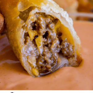 A hand dips a golden, crispy cheeseburger egg roll filled with juicy ground beef and melted cheese into a creamy orange sauce. Text below reads, Cheeseburger Egg Rolls - Crowd Favorite Appetizer.