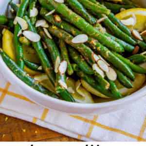 A white bowl filled with green beans, lemon slices, and almond slivers sits on a yellow and white towel, with whole and cut lemons in the background. Lemon Dijon Green Beans Almondine adds a zesty twist from The Food Charlatan.