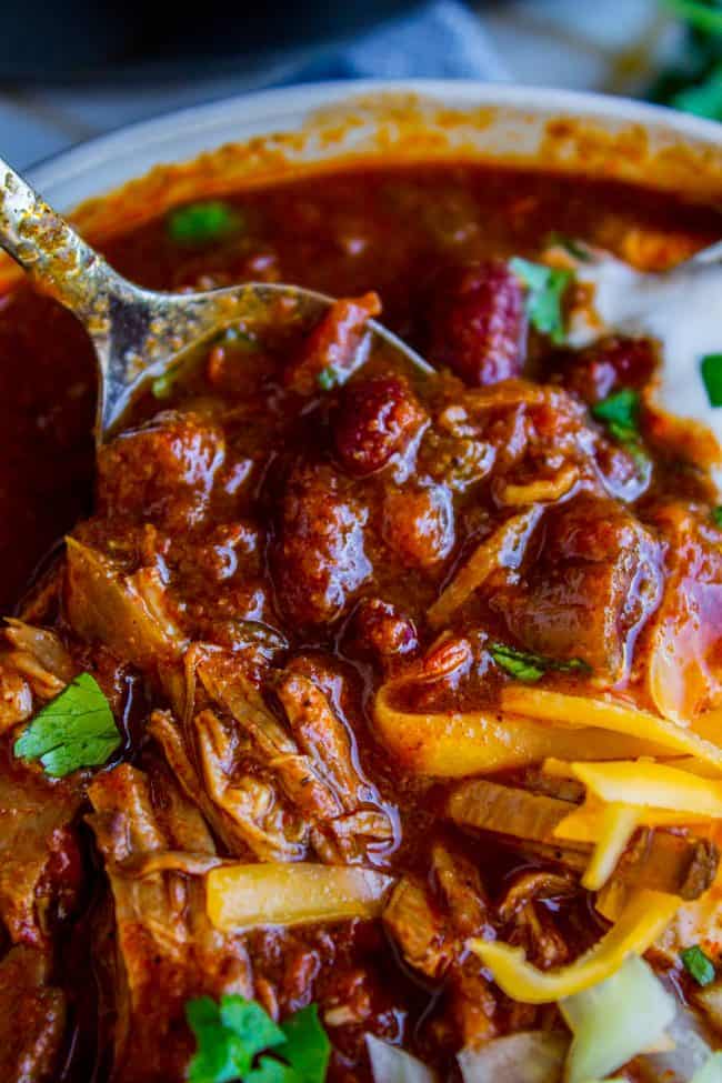 spoon being dipped into a bowl of chili topped with cheddar, onions, and cilantro.