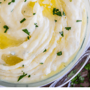 A close-up of creamy mashed potatoes in a glass bowl, topped with melted butter and sprinkled with chopped parsley. Text below reads: Classic Creamy Mashed Potatoes, The Food Charlatan.