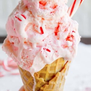 A hand holds a waffle cone filled with peppermint ice cream, swirled pink and white, topped with crushed candy canes and a small candy cane stick. Some of the peppermint ice cream is melting down the cone.
