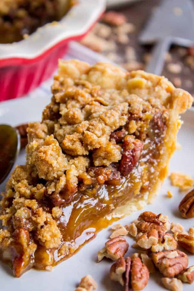A close-up of a slice of pecan pie with a crumbly topping, gooey filling, and whole pecans, set on a white plate with pecan pieces scattered around. The rest of the pie, inspired by a classic pecan pie recipe, is visible in the background.