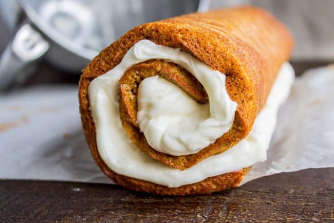 rolling a pumpkin roll with cream cheese frosting inside on a piece of parchment paper.