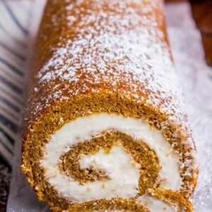 A pumpkin roll cake with a cream cheese filling is dusted with powdered sugar. One slice reveals the spiral pattern inside, showcasing this delightful pumpkin roll recipe on parchment paper, set against a softly blurred background.