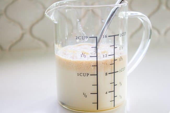 A glass measuring cup filled with a frothy light brown liquid and a metal spoon sits on a white surface against a tiled background. The cup is marked with measurements up to 2 cups or 16 ounces.