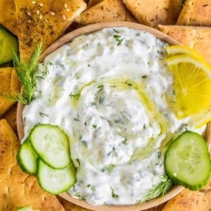 A bowl of creamy tzatziki sauce garnished with lemon slices, fresh dill, and cucumber, surrounded by pita chips, cucumber slices, and lemon wedges on a rustic wooden surface.