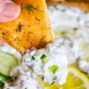 A hand holds a seasoned pita chip topped with creamy tzatziki sauce, garnished with herbs and green onions. Sliced lemon and cucumber add fresh color in the background.