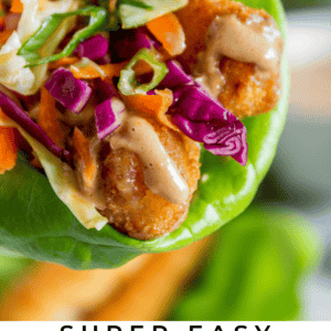A close-up of a hand holding a lettuce wrap filled with crispy fish, shredded carrots, red cabbage, cilantro, and sauce. Text below reads: Super Easy Thai Fusion Lettuce Wraps with Crispy Fish.