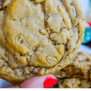 A hand with red nail polish holds a thick, chewy peanut butter oatmeal cookie. More cookies and a milk bottle are blurred in the background. Text reads: Melt in your mouth Peanut Butter Oatmeal Cookies. The Food Charlatan.