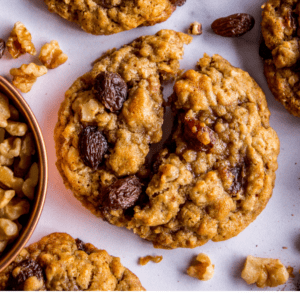 Close-up of soft oatmeal raisin cookies on a white surface, some whole and one broken in half, surrounded by raisins and chopped walnuts, with a text overlay reading The Very Best Oatmeal Raisin Cookies.