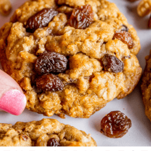 A close-up of a hand reaching for a soft, chewy oatmeal raisin cookie, with several cookies topped with plump raisins on a white surface. Text at the bottom reads, “The Very Soft and Chewy Oatmeal Raisin Cookies.”.