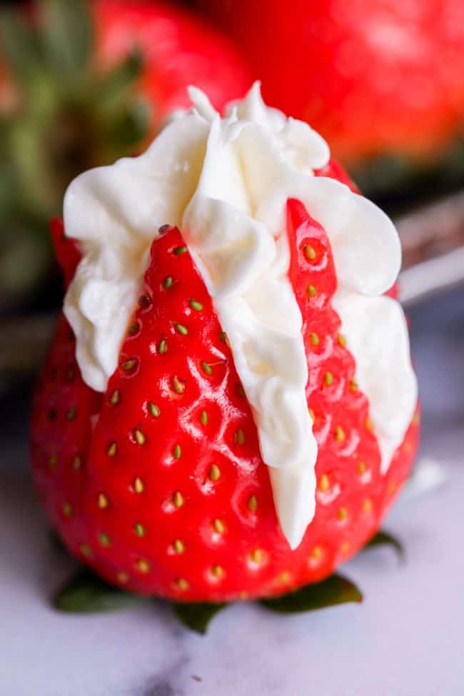 A fresh strawberry sliced partially open and filled with swirls of white whipped cream, creating cheesecake stuffed strawberries on a marble surface with other strawberries blurred in the background.