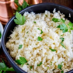 A black bowl filled with cauliflower rice and fresh parsley sits surrounded by parsley leaves and copper salt and pepper shakers on a wooden table.