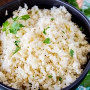 A black bowl filled with flavorful cauliflower rice garnished with fresh parsley sits on a patterned cloth, surrounded by herbs and salt and pepper shakers in the background.
