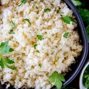 A black bowl filled with cauliflower rice, garnished with fresh parsley and surrounded by vibrant parsley leaves on a wooden surface.