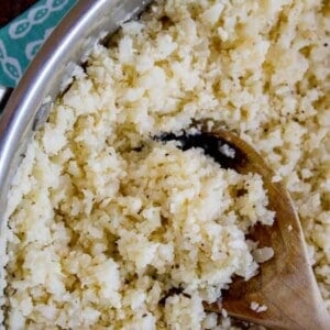 A close-up of cauliflower rice in a pan, being stirred with a wooden spoon.