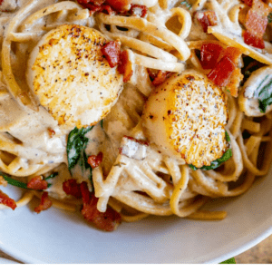A close-up of seared scallops on creamy pasta with spinach and bacon bits, topped with a rich champagne sauce. Text below reads: “How to cook scallops in creamy champagne sauce. The Food Charlatan.”.
