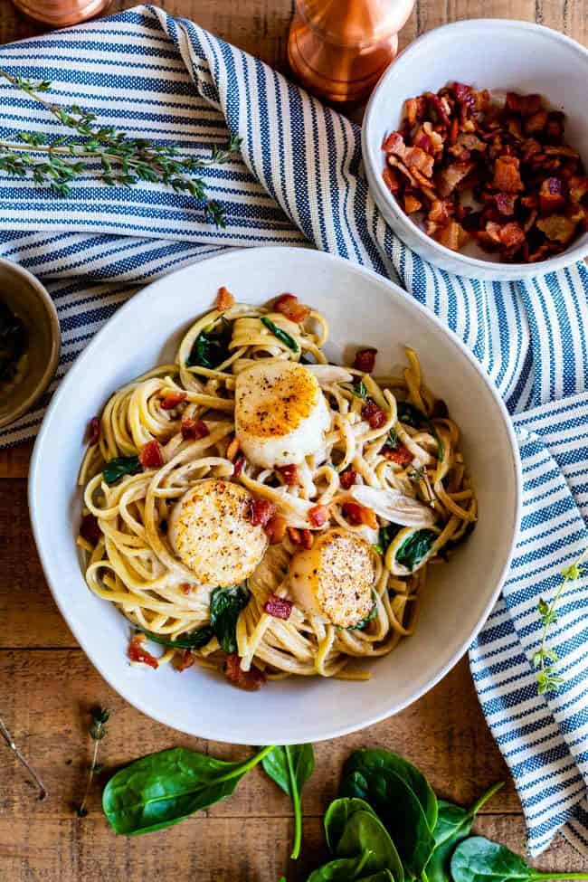 scallops with linguine, bacon, and spinach on a striped towel with a bowl of crumbled bacon.