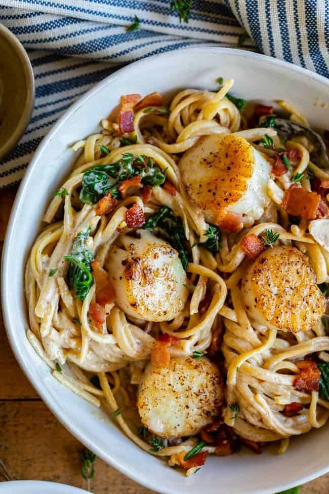 pan seared scallops with creamy wine sauce, bacon, and spinach over linguine in a white bowl. 