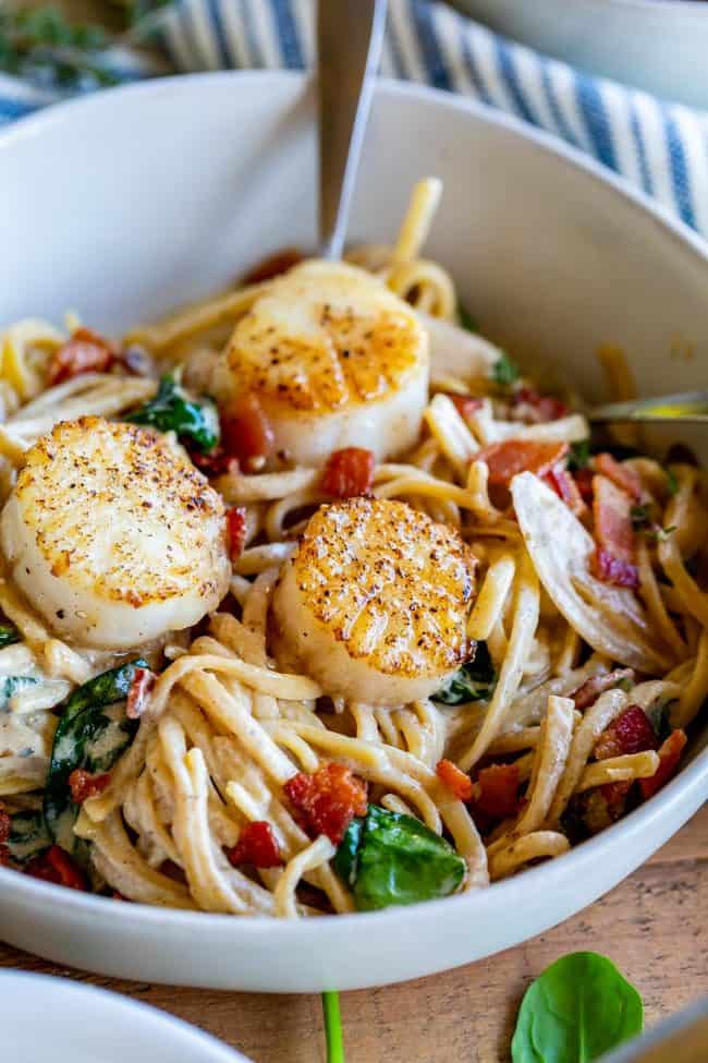 scallops with a creamy wine sauce, bacon, and spinach on linguine in a white bowl.