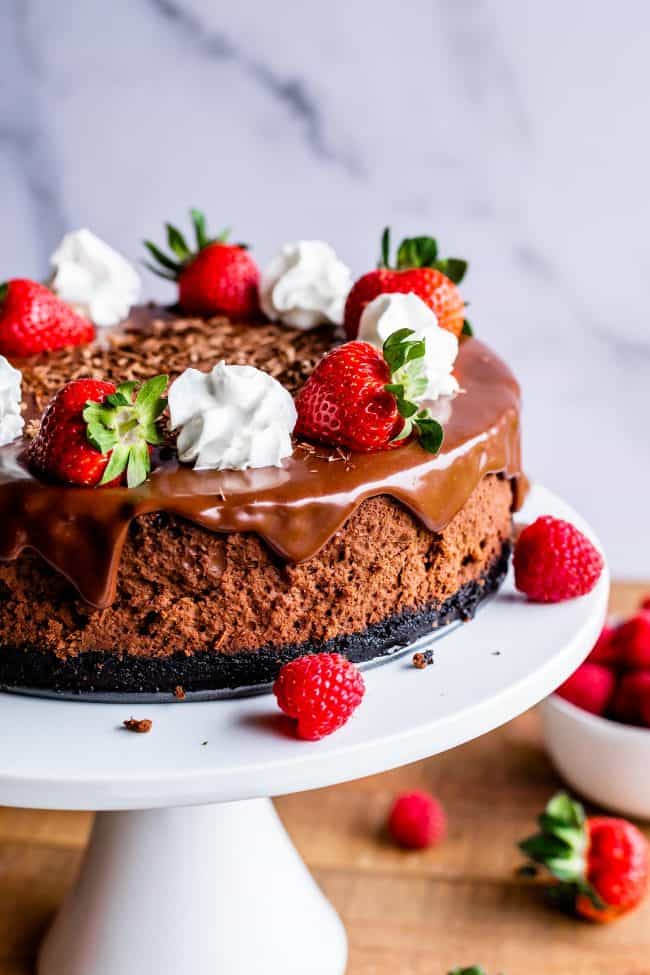 chocolate cheesecake with chocolate ganache, chocolate curls, whipped cream, and strawberries on a white cake stand.