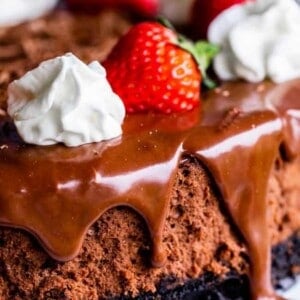 A close-up of a decadent chocolate cheesecake topped with chocolate ganache, fresh strawberries, and dollops of whipped cream. The luscious ganache drips down the side for an indulgent finish.