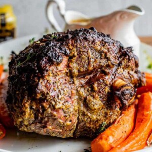 A roast leg of lamb with a crispy, seasoned crust is served on a white plate alongside roasted carrots. A white gravy boat is blurred in the background.