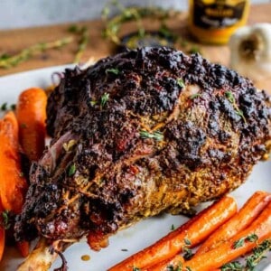 A roast leg of lamb with a dark, seasoned crust is served on a white platter, garnished with fresh herbs and surrounded by roasted carrots. A jar of mustard sits in the background.