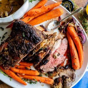 A platter with sliced roast beef and roast leg of lamb, glazed carrots, a bowl of brown gravy, and herbs. A spoon rests on the plate, with a jar of mustard, whole garlic, and a blue napkin nearby.