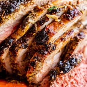 Close-up of a sliced, herb-crusted roast leg of lamb with a juicy, pink center, served alongside roasted carrots. The meat looks tender and flavorful with a crispy, seasoned outer layer.