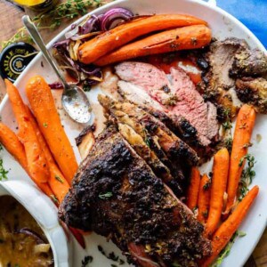 A platter with roasted beef ribs, sliced beef, whole cooked carrots, onion slices, a gravy boat, a jar of mustard, fresh herbs, a garlic bulb, and a flavorful roast leg of lamb beside a blue Walmart bag in the background.
