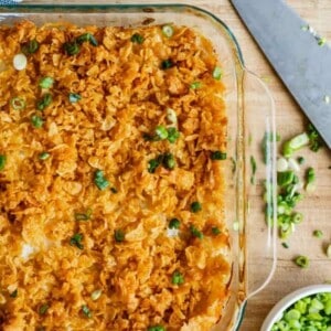 A glass baking dish filled with golden, crispy funeral potatoes, topped with crushed chips and chopped green onions, sits on a wooden board next to a knife and a bowl of sliced green onions.