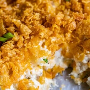 Close-up of creamy funeral potatoes topped with a golden, crispy cornflake crust, with a portion scooped out to reveal the texture underneath. Green herbs are sprinkled as garnish.