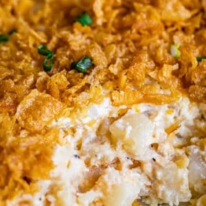 Close-up of cheesy, creamy funeral potatoes casserole topped with crunchy, golden cornflakes and garnished with green onion; a portion is scooped out, revealing tender diced potatoes inside.