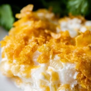 A close-up of cheesy funeral potatoes topped with a crispy golden cornflake crust and a garnish of fresh green herbs in the background.