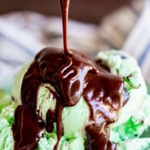 A close-up of mint chocolate chip ice cream in a bowl, with rich chocolate sauce being poured over the top.