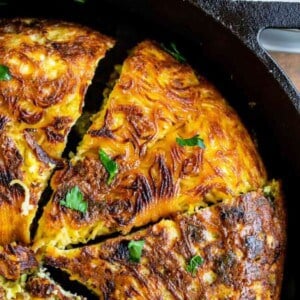 A close-up of a golden brown hash brown casserole, reminiscent of a pasta frittata, sliced into wedges in a cast iron skillet and garnished with fresh herbs. A portion sits on a white plate in the background.