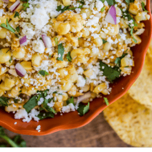 A bowl of Mexican street corn dip with cheese, cilantro, and red onion, surrounded by tortilla chips. The text below reads, Mexican Street Corn Dip - The Food Charlatan, featuring bold flavors and a festive touch.