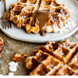 A plate of golden Liege waffles topped with crunchy pearl sugar, powdered sugar, and chocolate sauce sits on a table with a fork, napkin, and jar of sugar. Text reads: Classic Liege Waffles with Crunchy Pearl Sugar.