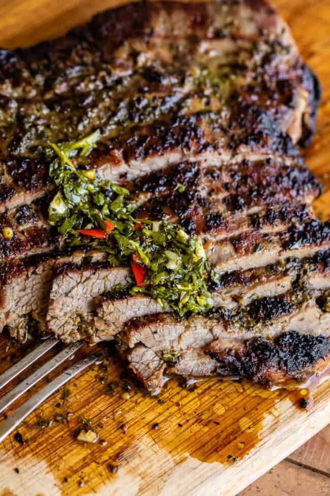 grilled sliced flank steak on a wooden cutting board with a metal fork, topped with chimichurri sauce.