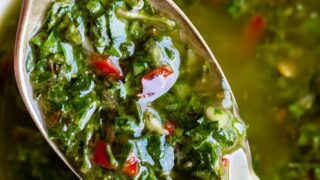 A close-up of a spoonful of vibrant chimichurri sauce, highlighting chopped parsley, garlic, red pepper flakes, and olive oil, with more chimichurri visible in a white bowl below.