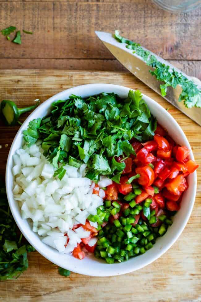 pico de gallo ingredients in a white bowl with a wooden cutting board and knife.