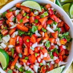 A bowl of fresh pico de gallo made with chopped tomatoes, onions, cilantro, and peppers, garnished with lime wedges, surrounded by tortilla chips and lime slices.