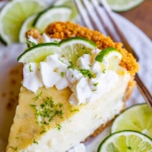 A slice of key lime pie topped with whipped cream and lime slices sits on a white plate, surrounded by extra lime slices and a fork. The pie features a crumbly crust and creamy key lime filling.