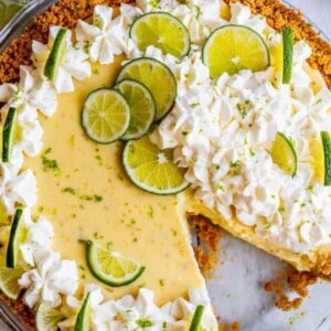 A key lime pie with a graham cracker crust, topped with whipped cream and fresh lime slices. One slice is missing, revealing the creamy key lime filling inside, while whipped cream and lime garnish decorate the top.