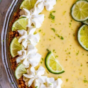 A close-up of a key lime pie with a graham cracker crust, topped with whipped cream swirls and fresh lime slices, displayed in a glass pie dish.