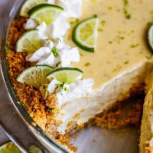 A close-up of key lime pie in a glass dish, topped with whipped cream and lime slices. A slice is missing, revealing the creamy filling and crumbly graham cracker crust, with lime wedges scattered nearby.