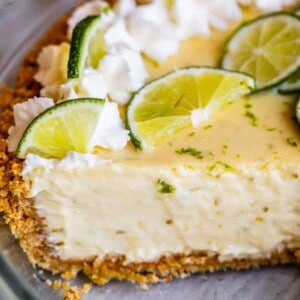 A close-up of a key lime pie with a graham cracker crust, creamy filling, and topped with whipped cream and fresh lime slices. A wedge has been cut from the pie, revealing its smooth texture.
